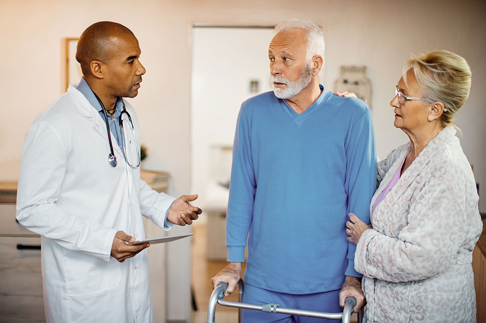 A doctor is talking to a senior couple. The man is using a walker and the woman is standing next to him with her hand on his shoulder.