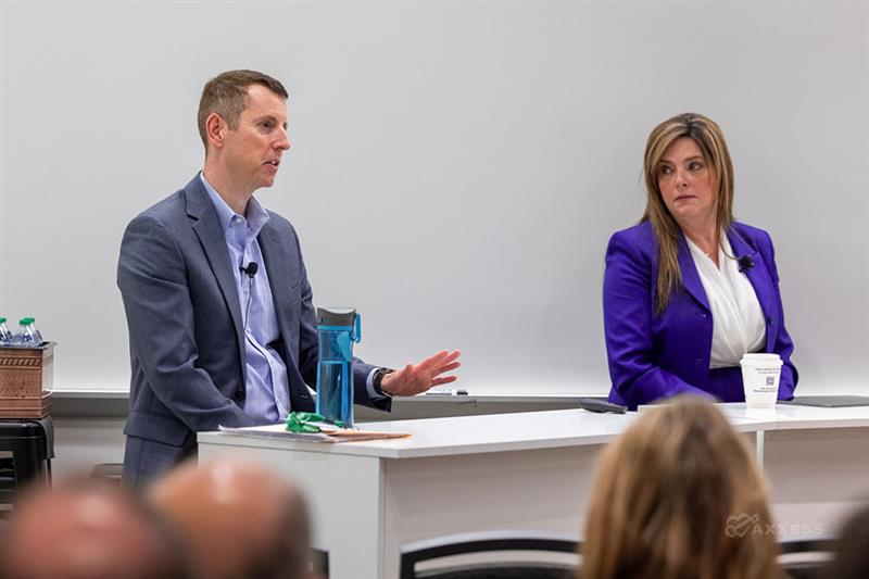 Two people, a man and a woman, are sitting at a table in front of a white board. The man is wearing a suit jacket and a blue shirt, and he is speaking. The woman is wearing a purple jacket and a white shirt, and she is listening. There is a water bottle on the table in front of the man, and a cup of coffee in front of the woman. The image is taken from the audience, and the heads of other people are visible in the foreground.