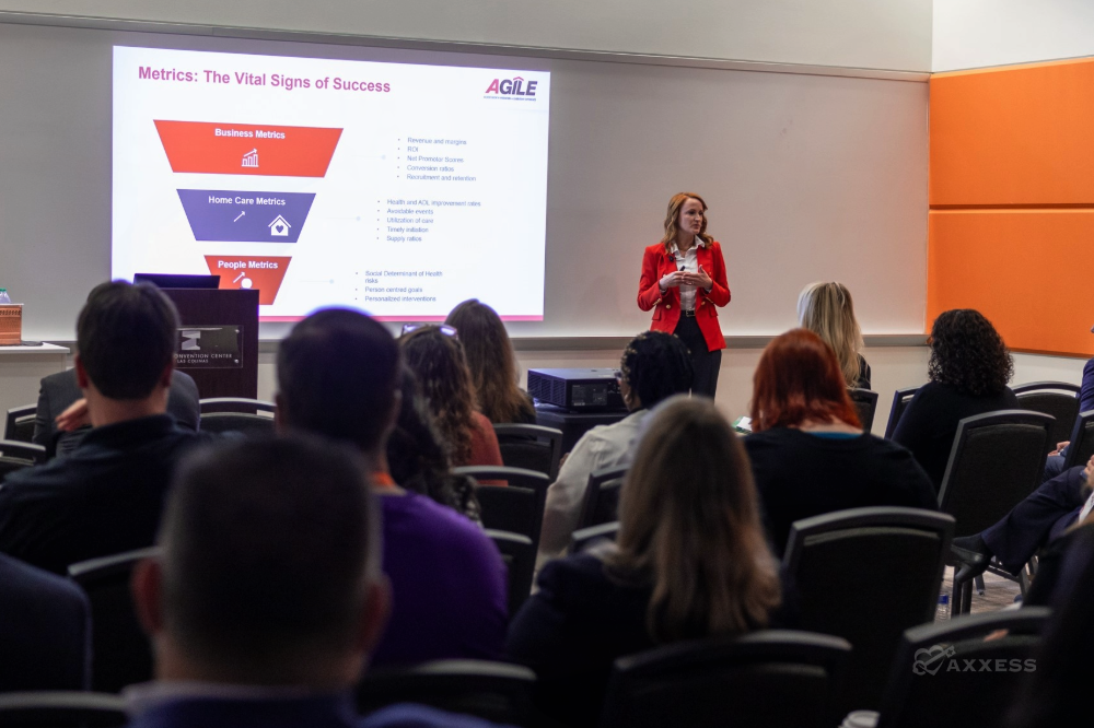 A woman in a red blazer presents a slide titled "Metrics: The Vital Signs of Success" in front of a large audience. The slide lists different metrics for the categories business, home care, and people. The room has an orange wall in the background.
