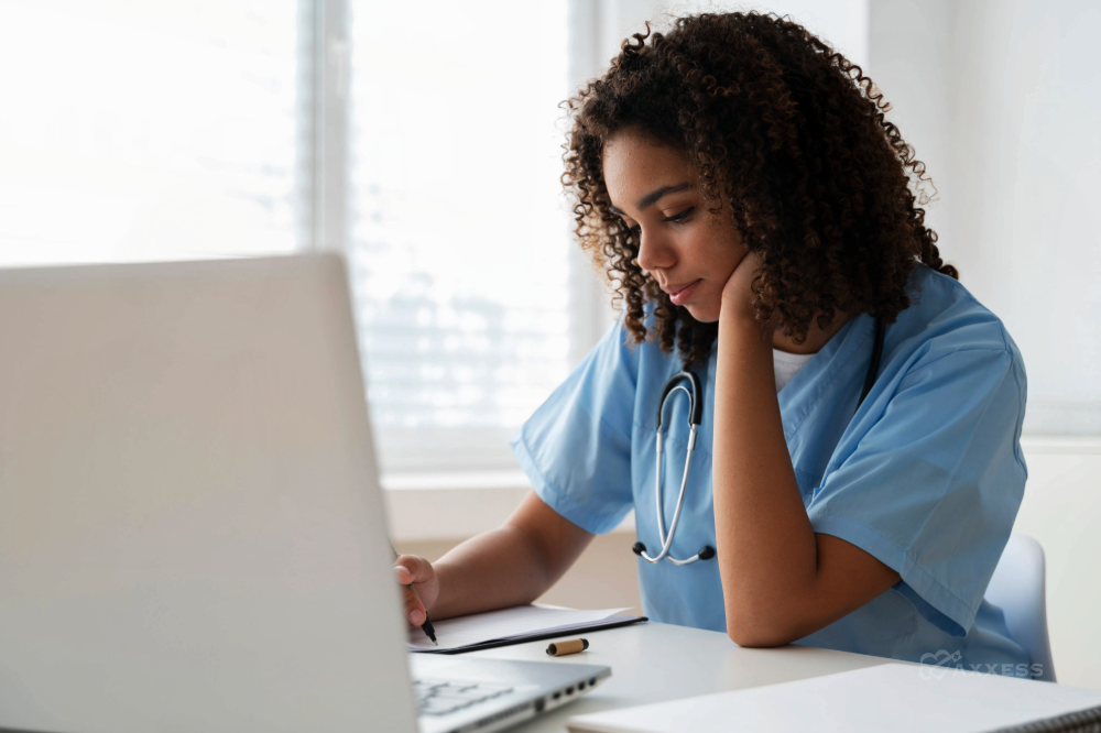 A young woman, wearing a blue medical scrub top and stethoscope, sits at a desk with a laptop and notepad, thoughtfully writing with a pen. She is a nurse diligently making notes. The white background emphasizes the focus on her work and the seriousness of the task at hand.