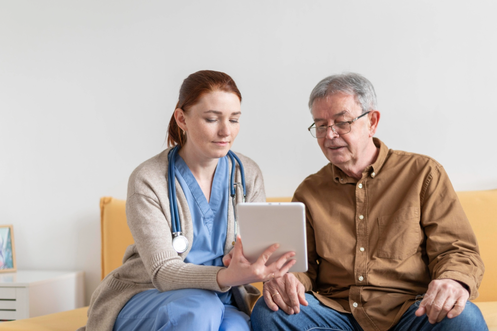 A female nurse in blue scrubs sits with a senior male patient on a yellow couch. She is holding a tablet and they are both looking at the screen.