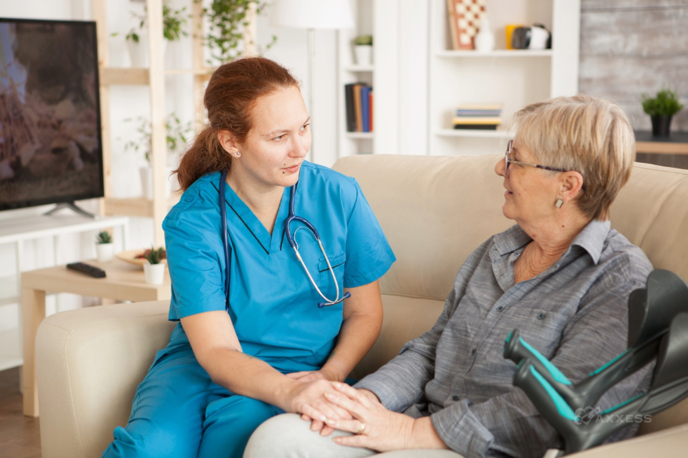 A young female nurse in blue scrubs is seated on a couch beside an elderly woman with an arm injury. The nurse is holding the woman's hand, and they are sharing a moment of connection.