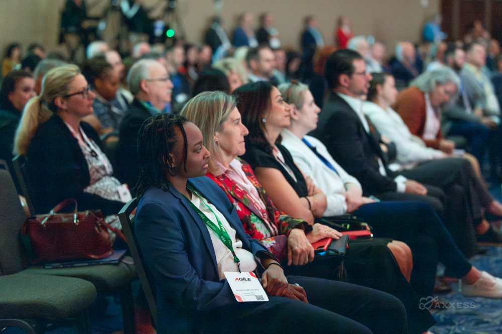 A diverse audience sits in rows of chairs at a conference. They are listening to a speaker who is out of frame. The image is cropped to highlight a woman in the foreground, who is listening intently with her arms crossed. She is wearing a blue blazer and has a name tag that reads "AGILE".